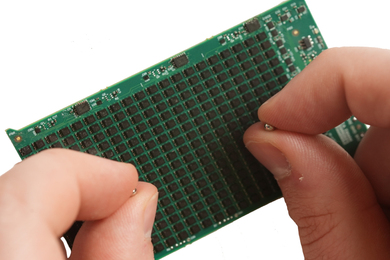 A hand holds two tiny beads over a green circuit board.