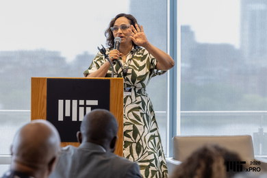 Prabha Jha speaks at a lectern in front of glass walls overlooking the Charles River