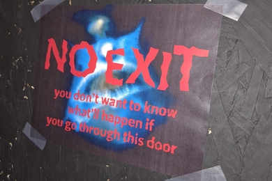 A sign with spooky font that reads: "No Exit - You don't want to know what'll happen if you go through this door" is taped to a black door
