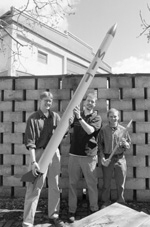 Graduate students Carl Dietrich (left) and Byron Stanley (center) and alumnus Andrew Heafitz with an actual-size model of the rocket they hope to launch, and a smaller prototype which will be launched in the next few weeks in preparation for November's contest.