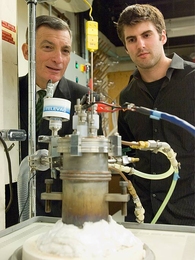 Professor Donald Sadoway and graduate student David Bradwell observe one of their small test batteries in the lab. The battery itself is inside the heavily insulated metal cylinder at center, which heats it to 700 degrees Celsius, while the wires at top charge up the battery and measure its performance.