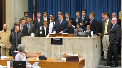 About 17 members of MIT's Men's Basketball team and middle-school aged student athlete surround podium.
