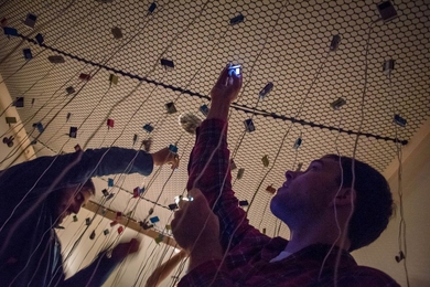 A man holds a phone up to a net overhead to take a photo. Strings hang down from the net.