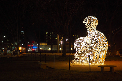 Photo of a white sculpture on the MIT campus, shaped like a person sitting, lit up at night