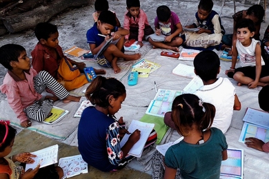 Photo of about a dozen young children sitting on the ground doing schoolwork.