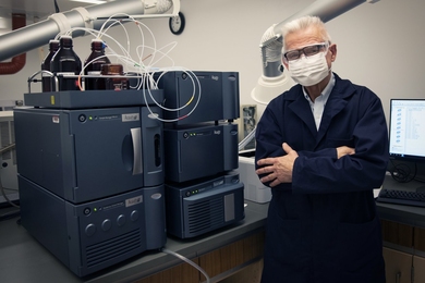 Photo of masked John Dolhun standing next to a new piece of lab equipement that looks like a stack of computers attached to bottles of liquid on top