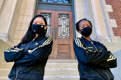 Photo of Jeana Choi and Koumani Ntowe in black sweatshirts and black face masks, posing with arms crossed in front of an MIT building.