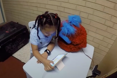 Photo of a young girl sitting at desk doing homework on a tablet next to a fluffy orange and blue robot.