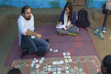 Photo of a man and a woman sitting on a rug with cards laid in front of them. The man is talking and the woman is writing in a notebook.