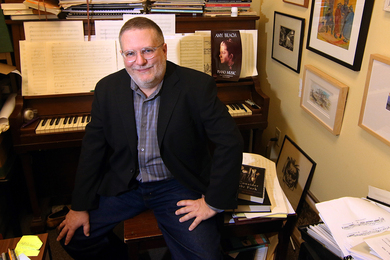 Photo of MIT composer Charles Shadle sitting at his piano, facing the camera and smiling