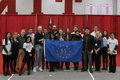 Photo of 21 young people standing together in front of red curtain divider in an MIT gym and holding a modified European Union flag with a circle of stars and "mens" and "manus" personified inside the stars
