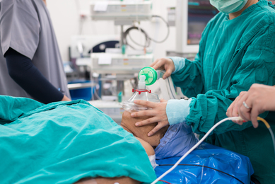 A sedated patient in an operating room receives attention from an anesthesiologist, who holds a gas mask over their mouth and nose