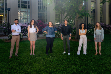 Photo of six people standing in a line, with spring foliage and an MIT building in the background