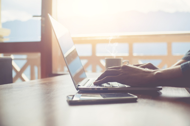 Photo of a person's hands working on a laptop computer that's on a table next to a balcony