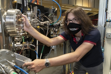 Photo of Jonathan Pelliciari in a lab, wearing a face mask and holding onto a large instrument with many metal knobs and wires