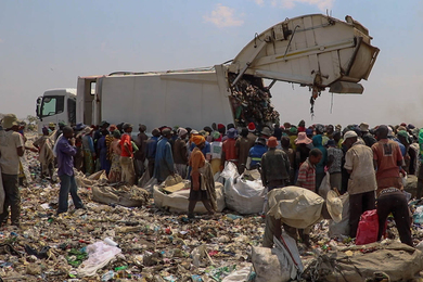A crowd of people surround a truck unloading waste at a landfill site