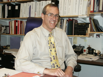 Man in a tan shirt, yellow tie, and glasses is smiling at his in front of a crowded book case 