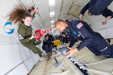 Photo of a man in a blue jumpsuit operating a handheld device while floating inside an airplane cabin; a woman in a green jumpsuit watches, while several others use various devices of their own in the background.