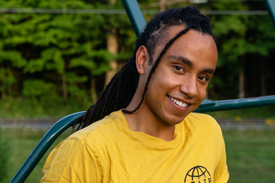 Miles Johnson, with long braids and a bright yellow t-shirt, smiles while leaning on what looks like metal playground equipment in a park