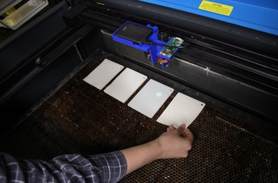 Photo of a hand holding a flat white square that is being printed-upon by a computer-printer-like machine that is open so you can see it carving shapes into the white material