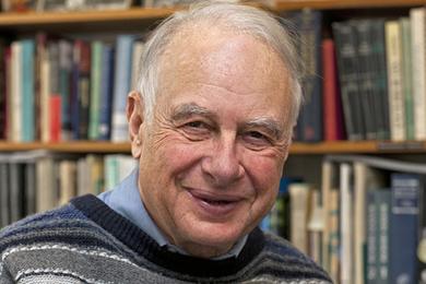 Headshot photo of a smiling man wearing a striped sweater and standing in front of book shelves