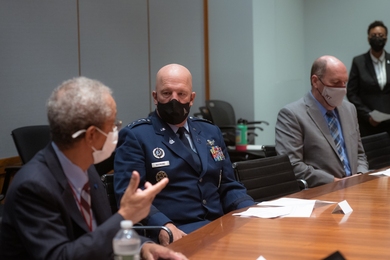Photo of three men wearing facemasks and sitting at a table, with another indivdiual standing in the background