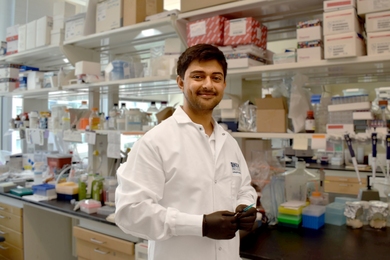 Photo of Sachin Bhagchandani standing in a lab, wearing a white coat and black safety gloves