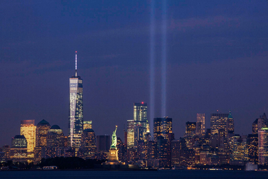 Photo of the New York City downtown skyline taken at dusk, with the Statue of Liberty in the foreground and two large blue beams of lights emerging from where the World Trade Center used to stand
