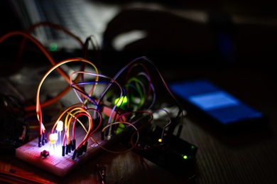 Photo of a circuit board on a tabletop, with the room lights turned out. The circuits are lit with two small LEDs in pink and green.