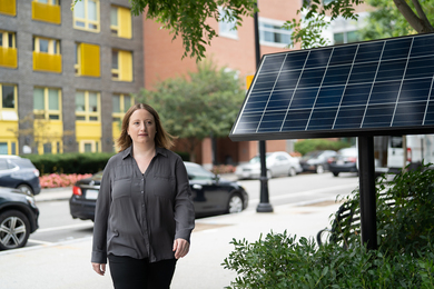 Photo of Noelle Eckley Selin walking down a street with brick buildings in the background and a large solar cell panel atop an approximately 2-meter-tall pole