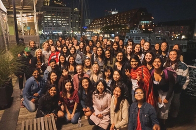 Group photo of approximately 50 women taken at night with a cityscape as well as a boat and water in the background.