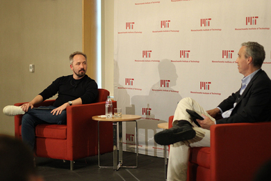 Photo of Drew Houston and Dan Huttenlocher, seated in red chairs and speaking to each other in front of a wall with a repeating MIT logo