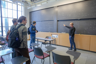 Photo of George Barbastathis standing at a chalkboard, gesturning and explaining something to three standing students