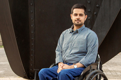 Photo of Rijul Kochhar seated in a wheelchair, in front of large sculpture