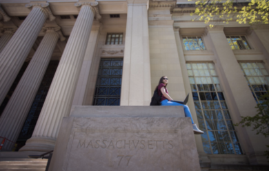A student sits with a laptop open on a stone slab in front of two MIT buildings. The slab says "Massacvsetts 77"