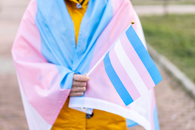 A person is wrapped in the transgender pride flag and is holding a small transgender pride flag.