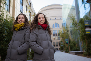Photo of nearly-identical twin sisters posing back-to-back in matching winter jackets in the courtyard in between MIT Building 10, with a stone facade, and MIT.nano, with an all-glass facade.