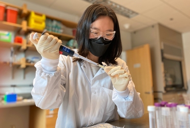 Photo of Anna Yeh wearing goggles, a lab coat, a mask, and gloves and uses a pipette in a lab