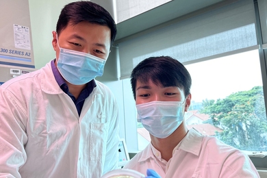 Photo of two fully gowned researchers standing in a lab, in front of a window with trees in the background