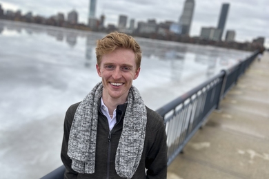 Portrait of Alex Tinguely on a bridge with Boston behind him. It’s winter, and the river is frozen