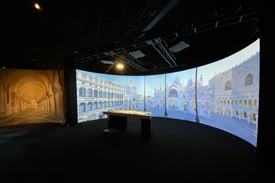 Photo of a large, dark room. There is a semi-circular screen display, showing St. Mark’s Basilica in Venice. In the middle of the room is a table.