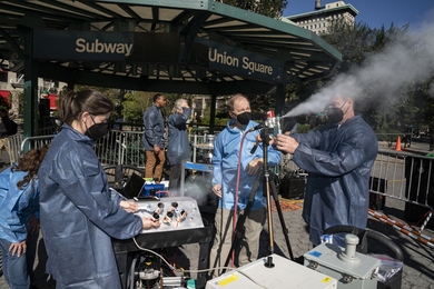 Photo of six scientists in blue lab coats standing under or near a metal gazebo that says "Subway, Union Square." Two of the scientists operate a small sprayer, on a tripod, that is shooting out what looks like mist. 