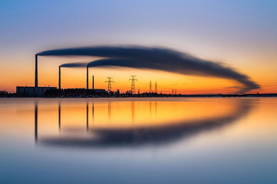 three smokestacks billow symmetric plumes of smoke over the horizon, which is reflected in a pool of still water below.