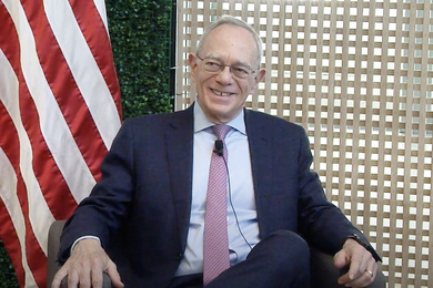 L. Rafael Reif, smiling, sits in front of a wooden screen and a U.S. flag