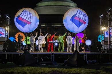 Photo of a group in colorful costumes with their backs to the camera and right hands pointed to the right with the MIT Dome in the background.