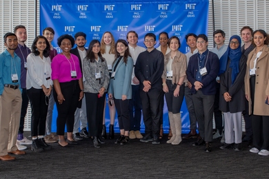 Photo of a large group of students standing together and posing in front of a blue banner with "MIT PKG Ideas" logos