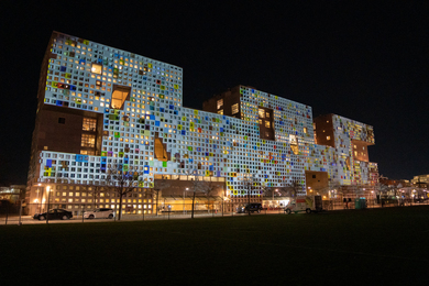 Photo of a multicolored art installation at night on the facade of Simmons Hall, a rectangular dorm with a brushed metallic gray facade, half a dozen large cutouts, and many small square windows