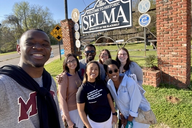 Photo of seven students posing in front of a large road sign that says "Welcome to historic Selma, queen city of the blackbelt"