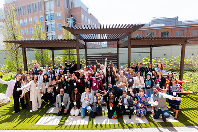 About 50 Solve at MIT attendees pose for a group photo. They are posing with arms out in all directions and standing on grass in front of a wooden platform, which is next to a couple of brick buildings