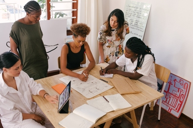 Photo of five women surrounding a table during a meeting. One works on a tablet, two write on paper, and the last two look at their colleagues' work.